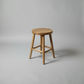 A natural wood stool with a round seat and four angled legs, shown on a neutral gray studio background.