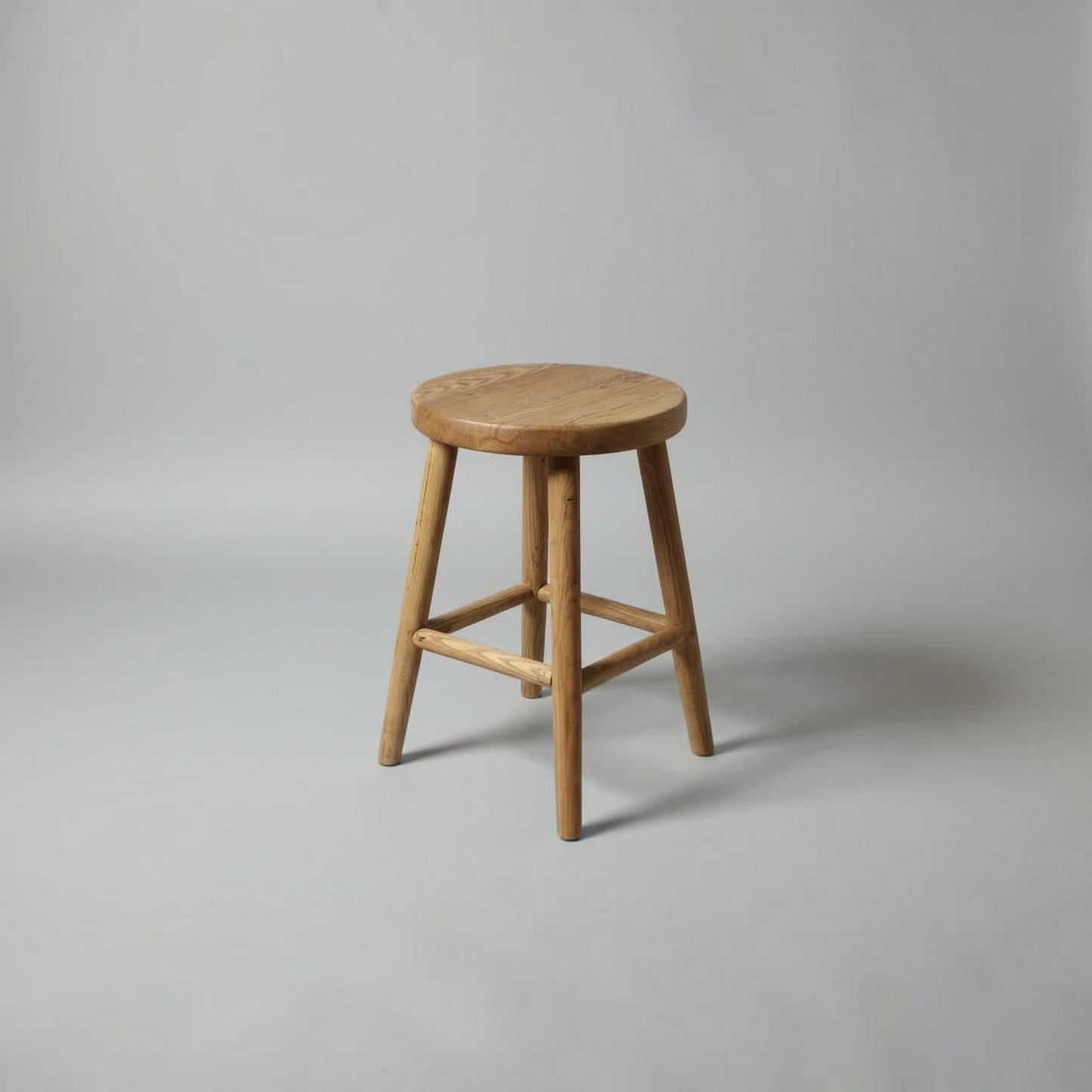 A natural wood stool with a round seat and four angled legs, shown on a neutral gray studio background.