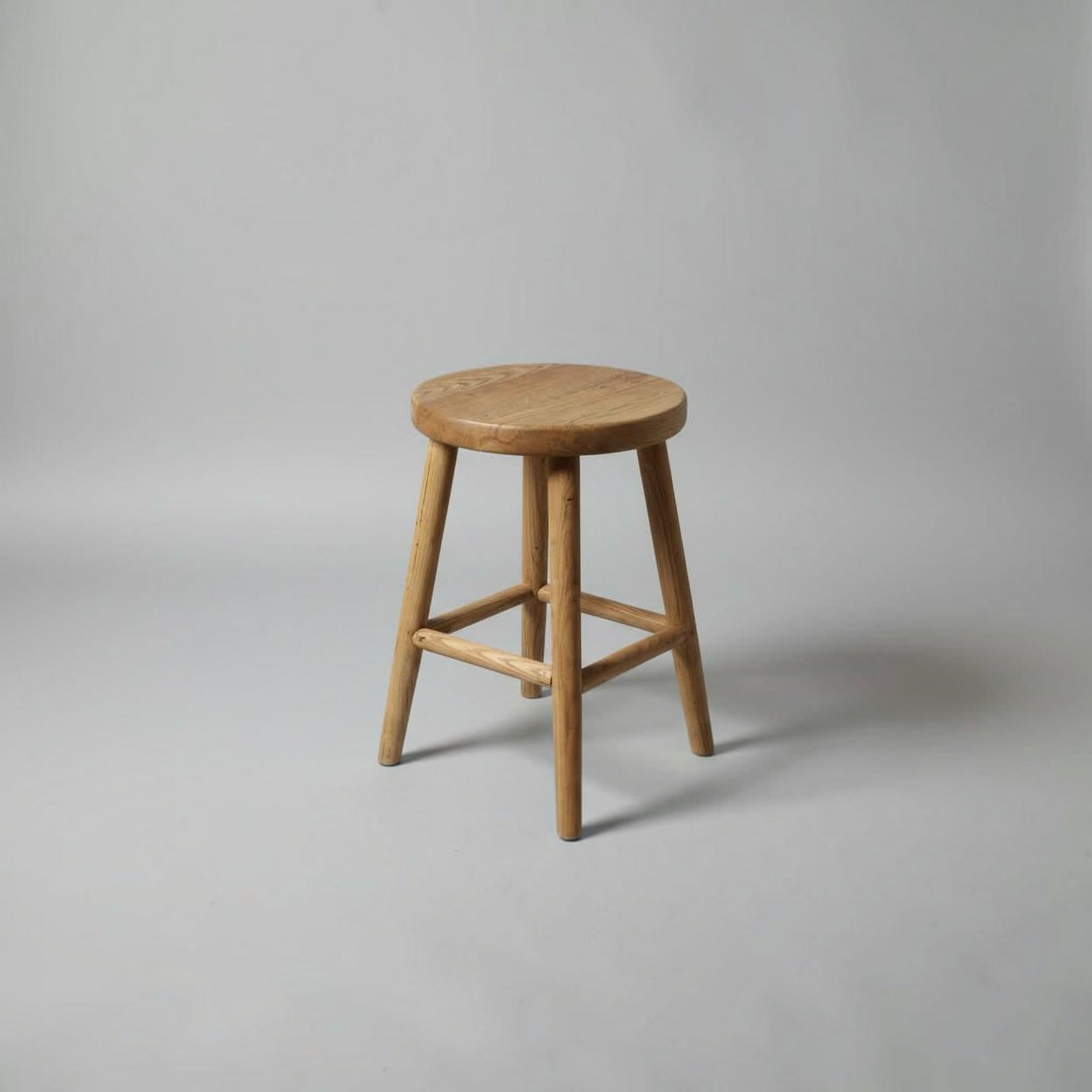 A natural wood stool with a round seat and four angled legs, shown on a neutral gray studio background.