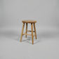 A rustic wooden stool with a round seat and four angled legs, shown against a neutral gray studio background.