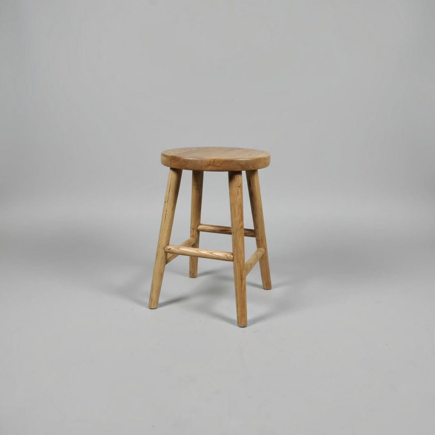 A rustic wooden stool with a round seat and four angled legs, shown against a neutral gray studio background.