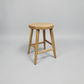 A light natural wood stool with a round seat and four angled legs, shown on a neutral light gray background.