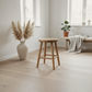 A light natural wood stool with a round seat and four legs stands in a bright, minimalist room with pale wood floors, a large white vase with pampas grass, and a bench in the background.