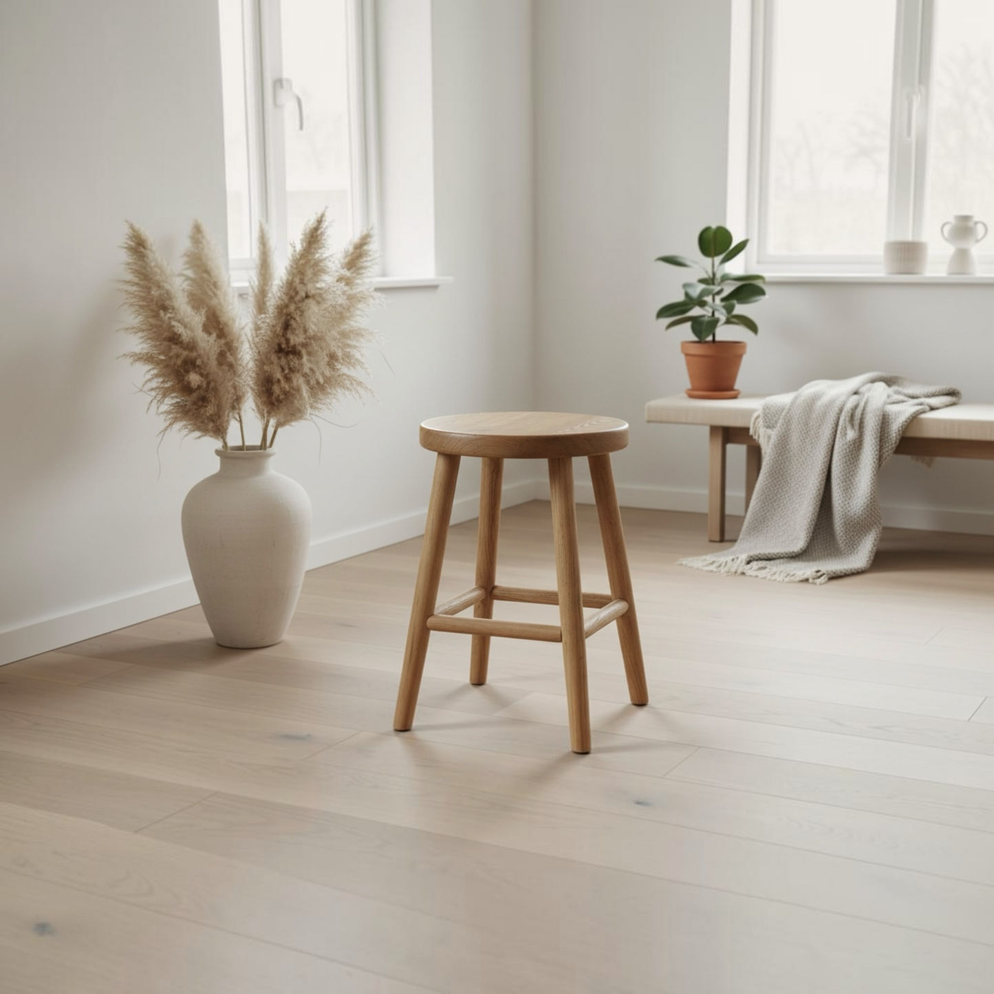 A light natural wood stool with a round seat and four legs stands in a bright, minimalist room with pale wood floors, a large white vase with pampas grass, and a bench in the background.