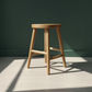 A light natural wood stool with a round seat and four angled legs, positioned on a light wood floor against a dark green wall with soft sunlight and shadows.