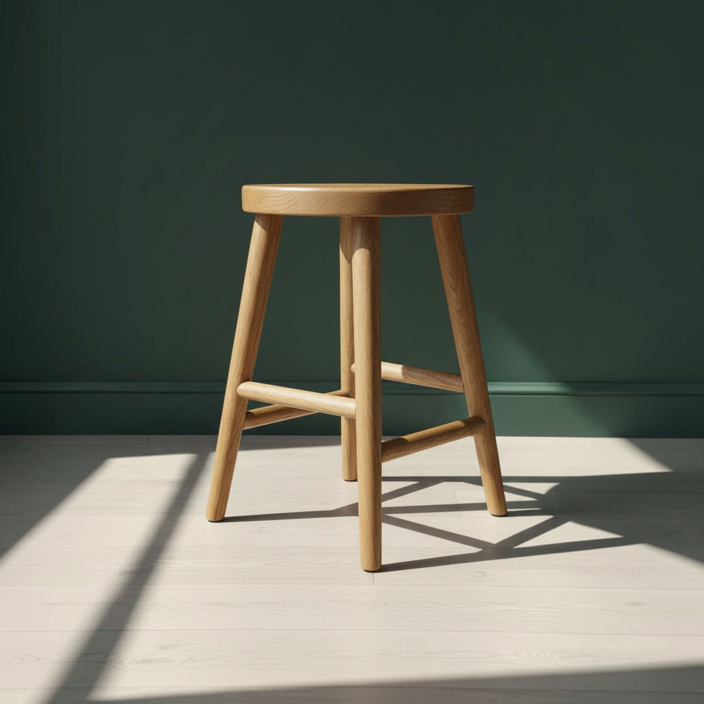 A light natural wood stool with a round seat and four angled legs, positioned on a light wood floor against a dark green wall with soft sunlight and shadows.