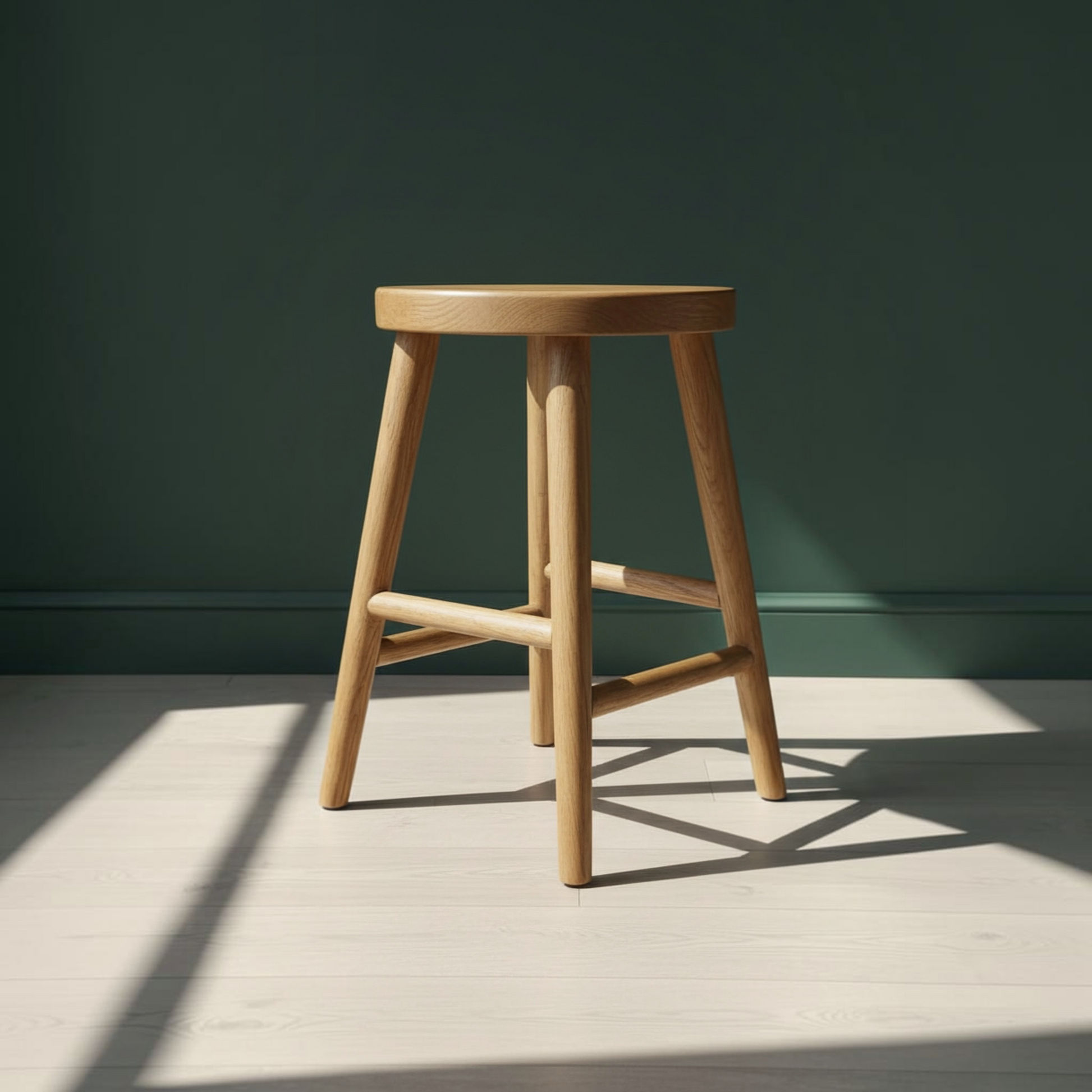 A light natural wood stool with a round seat and four angled legs, positioned on a light wood floor against a dark green wall with soft sunlight and shadows.