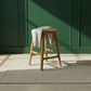 A light natural wood stool with a draped off-white throw blanket, set on a textured cream rug against a dark green paneled wall with soft sunlight casting shadows.