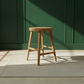 A round wooden stool with four legs and crossed front support bars, shown on a light rug against a dark green paneled wall with sunlight casting shadows.