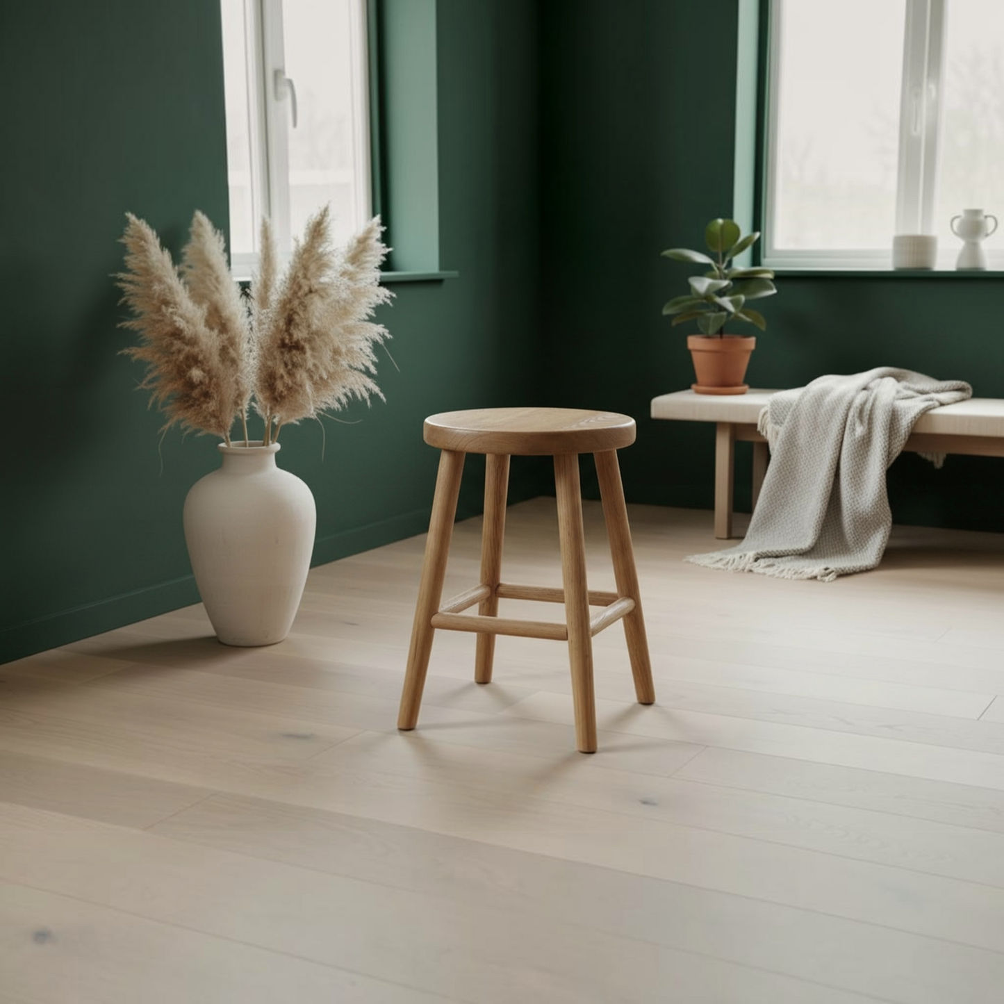 A light natural wood stool with a round seat and four legs stands in a minimalist room with dark green walls, light wood flooring, a white vase with pampas grass, and a bench with a throw blanket.