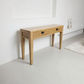 A light oak wood console table with two drawers and black metal handles, set against a minimalist white wall in a bright room.