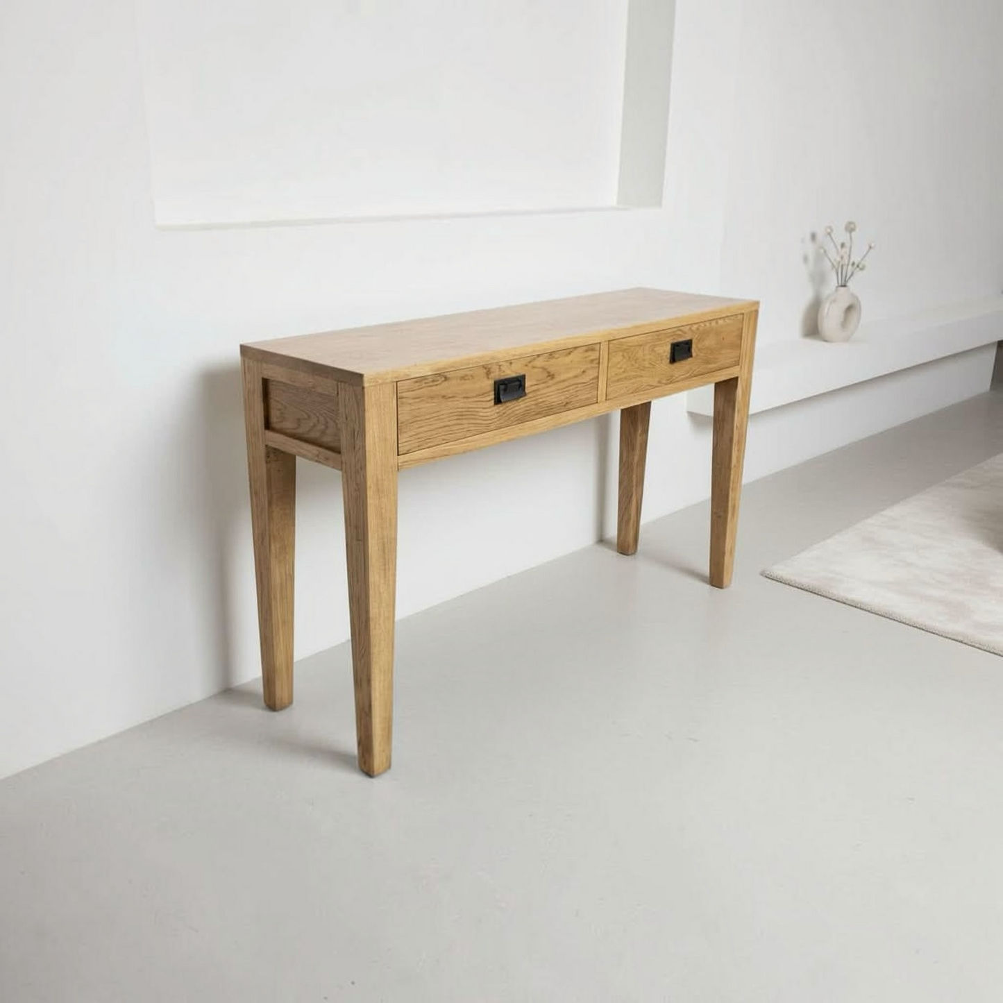 A light oak wood console table with two drawers and black metal handles, set against a minimalist white wall in a bright room.
