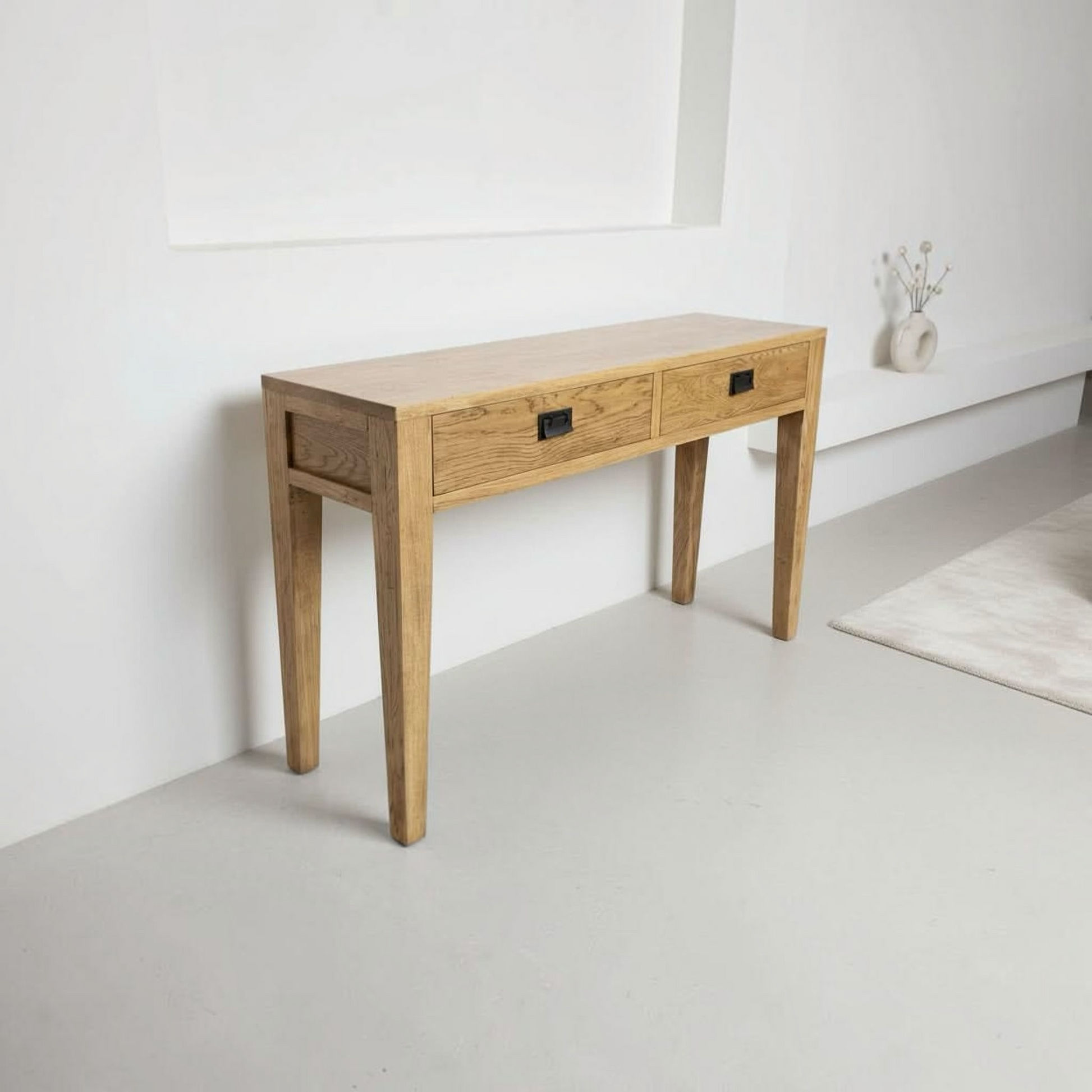 A light oak wood console table with two drawers and black metal handles, set against a minimalist white wall in a bright room.