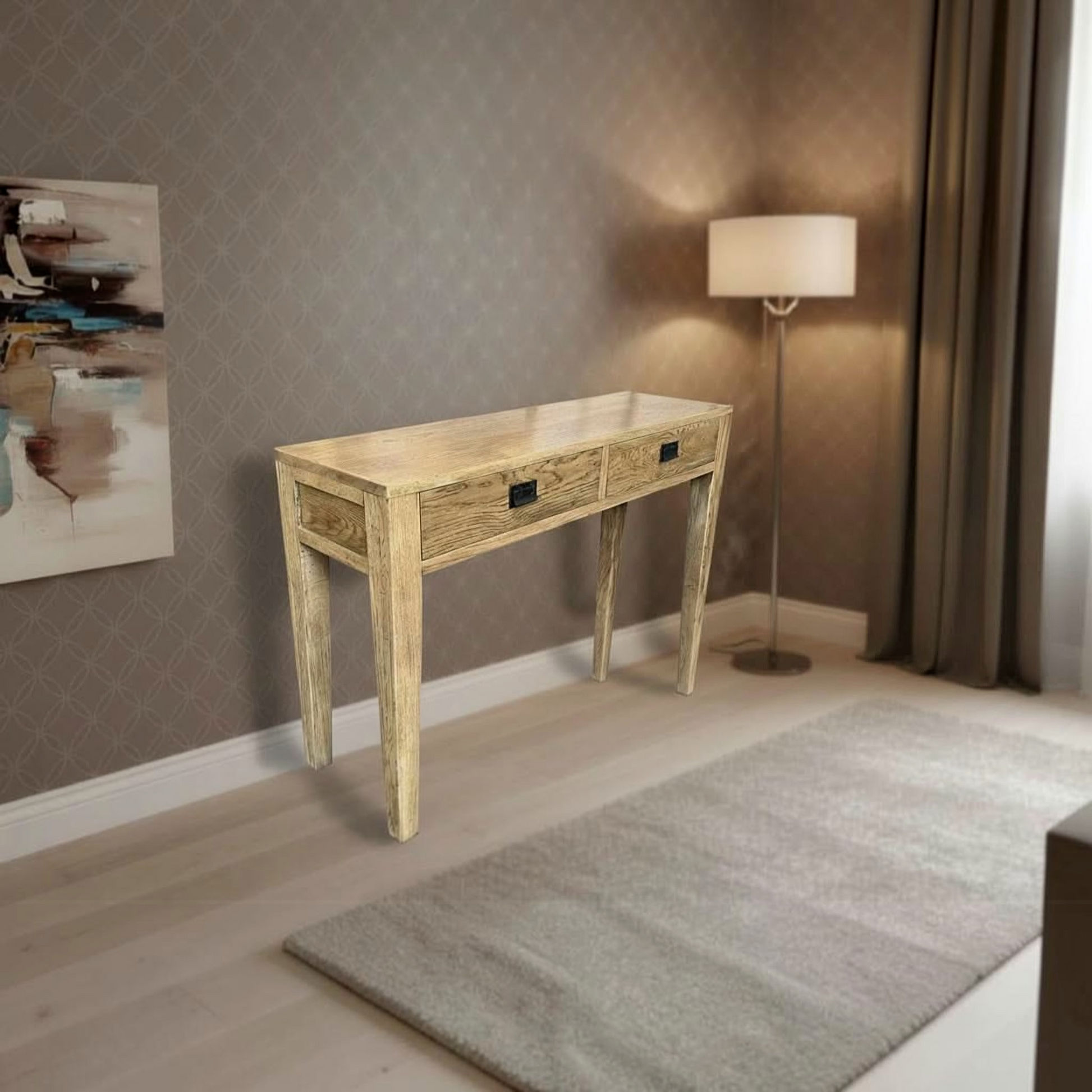 A rustic light wood console table with two drawers and black metal handles, styled in a modern living room setting with a gray rug, floor lamp, and patterned wallpaper.