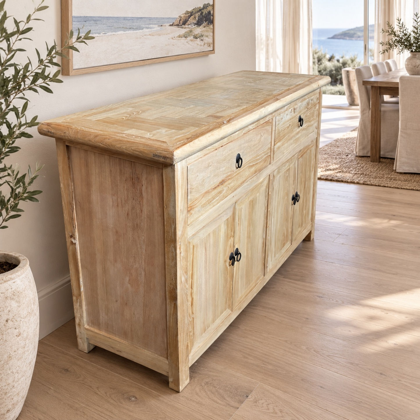 A light natural wood storage cabinet with two top drawers and four lower cabinet doors, styled in a bright, modern living room with light wood floors and a large window.