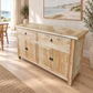 A light natural wood sideboard or buffet cabinet with two drawers on top and three cabinet doors below, shown in a bright, modern room with a beach painting on the wall and a window in the background.