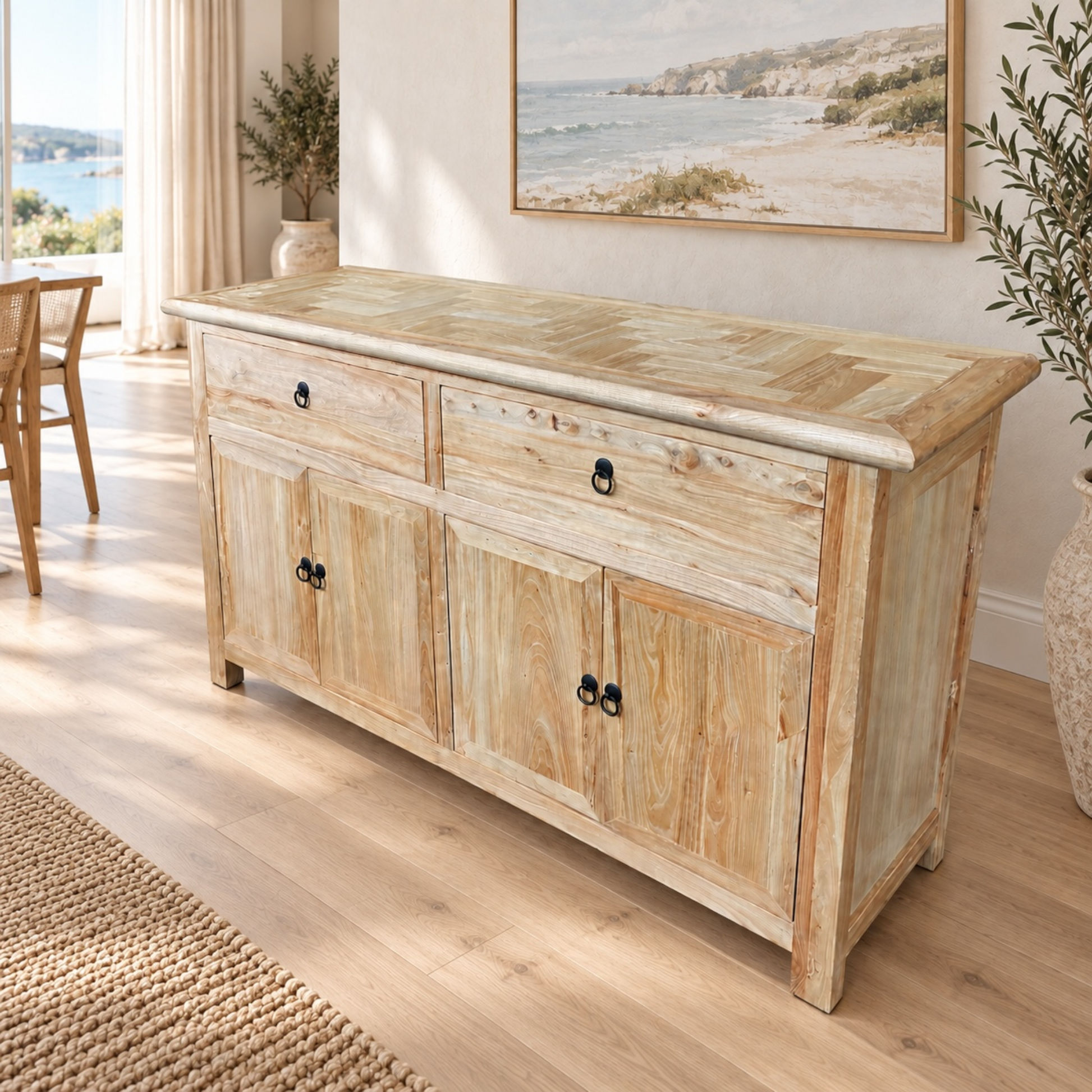 A light natural wood sideboard or buffet cabinet with two drawers on top and three cabinet doors below, shown in a bright, modern room with a beach painting on the wall and a window in the background.