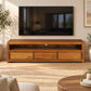 A warm medium-brown wooden TV stand with three drawers and open shelving, shown in a styled living room setting under a wall-mounted television.