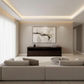 A modern living room with a large abstract art print hanging on a beige wall above a dark wood console table. The room features a light beige sectional sofa, a round glass coffee table, and a white shag rug, with soft natural light coming through sheer curtains on the left.