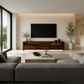 A modern living room with a large black flat-screen TV mounted on a beige wall above a dark wood media console. The room has a low-profile gray sectional sofa in the foreground, a light stone coffee table, and tall potted plants on either side of the TV.