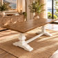 A rustic farmhouse dining table with a medium brown wood top and white trestle-style legs, shown in a bright, modern dining room setting on a woven jute rug.