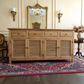 A light natural wood sideboard or credenza with four louvered cabinet doors and three drawers, styled in a classic room setting on a patterned rug with a gold mirror and sconces behind it.