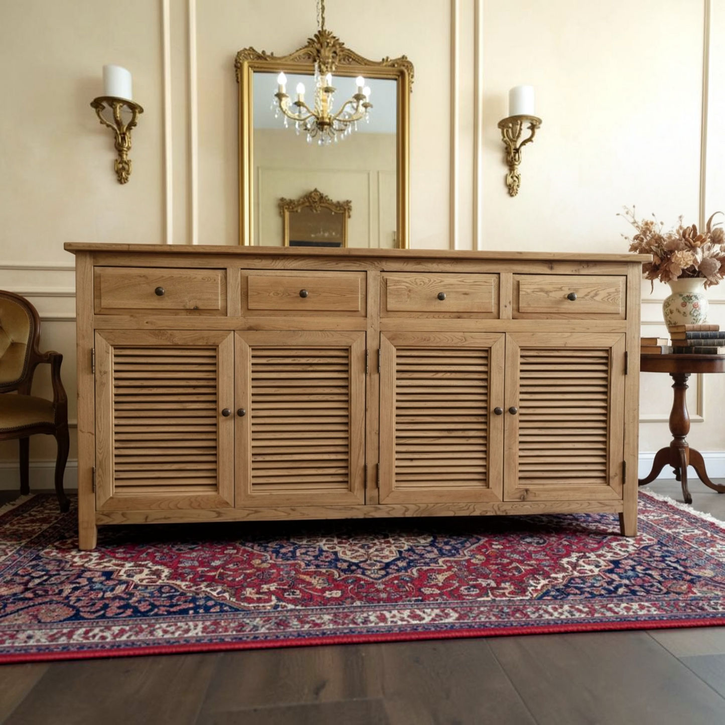A light natural wood sideboard or credenza with four louvered cabinet doors and three drawers, styled in a classic room setting on a patterned rug with a gold mirror and sconces behind it.