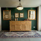 A light natural wood louvered sideboard cabinet styled in a home interior with a dark green wall, gold-framed mirrors, a hanging pendant light, and a patterned area rug.