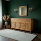 A light natural wood sideboard cabinet with louvered doors and drawers, styled in a dark green room under a gold-framed mirror and wall sconces.
