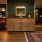 A rustic wooden sideboard cabinet with louvered doors and three drawers, styled in a traditional room with a gold mirror, books, and a rug.