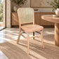 A light natural wood dining chair with a woven cane backrest, positioned on a textured beige rug in a bright, modern dining room next to a round wooden table.