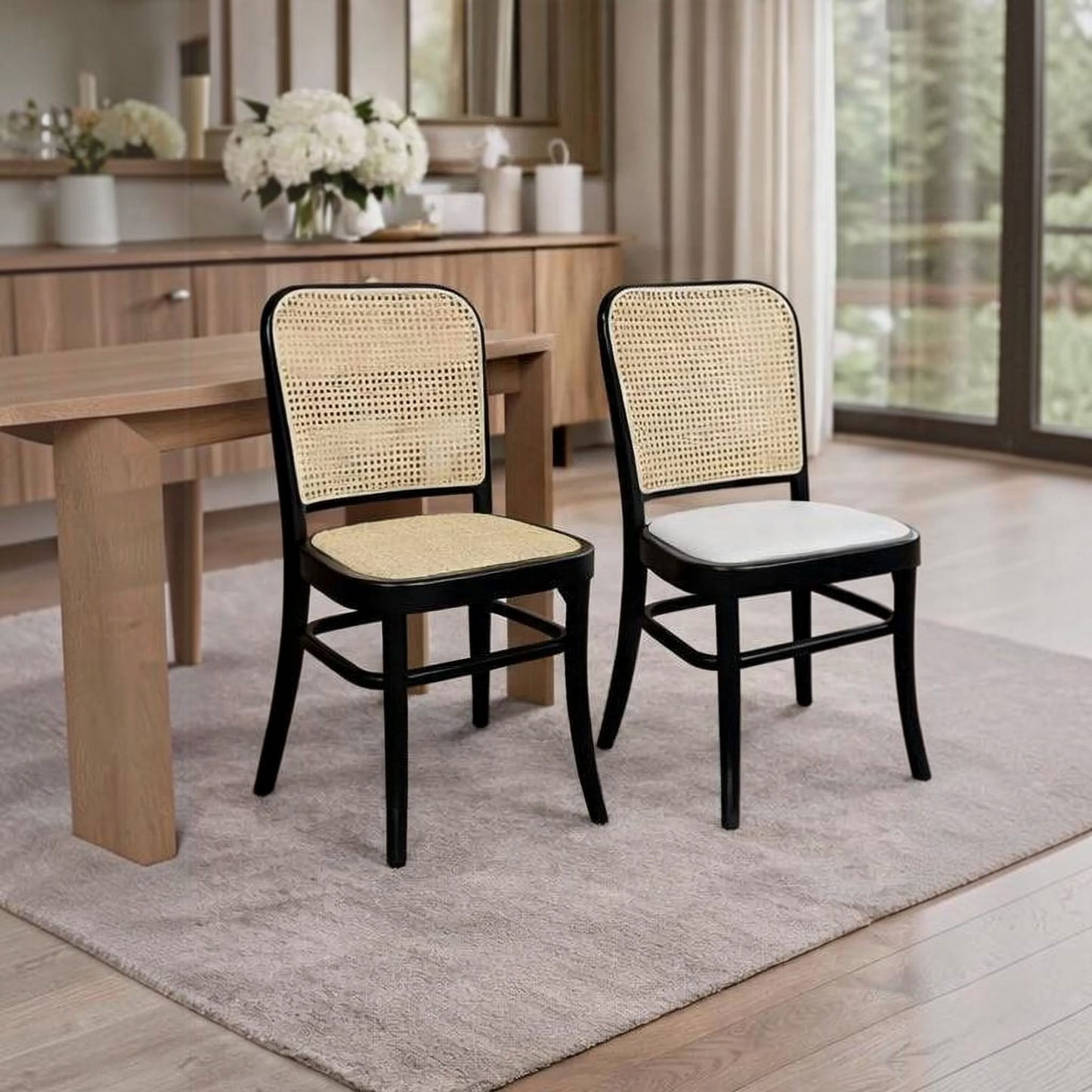 Two dining chairs with black frames and woven cane backs, one with a light wood seat and one with a white seat, placed in a bright dining room next to a wooden table.