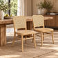 Two light natural wood dining chairs with woven cane backs are placed in front of a rustic wooden dining table in a warm, sunlit room with a rug, plants, and curtains.