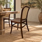 A dark brown wooden dining chair with a woven cane backrest, shown in a styled dining room setting next to a rustic wooden table on a woven jute rug.