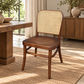 A wooden dining chair with a woven cane backrest, positioned in a bright, modern living space next to a rustic wooden table on a textured beige rug.