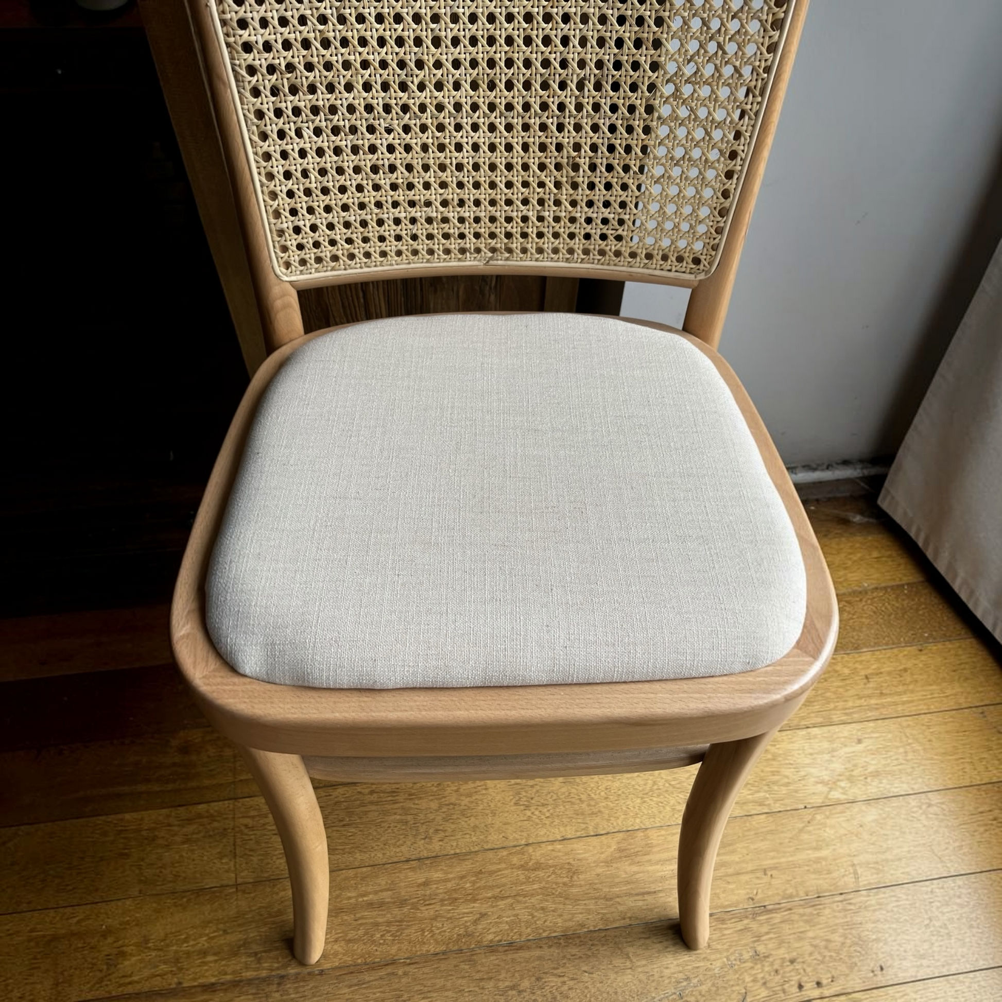 A wooden dining chair with a woven cane backrest and a light beige upholstered seat cushion, shown in a home setting on a wooden floor.