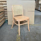 A light natural wood dining chair with a woven cane backrest, positioned in front of a large cardboard panel on a concrete warehouse floor.