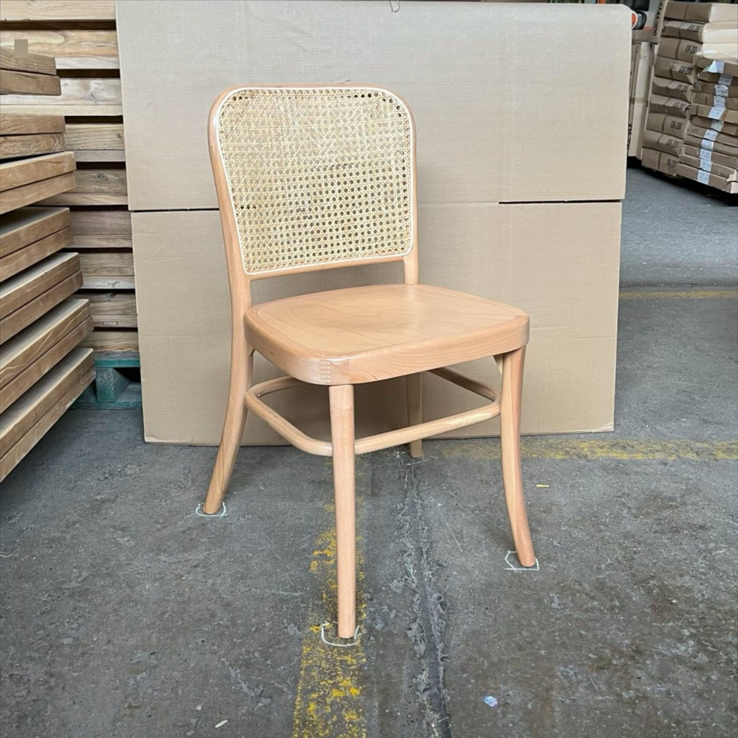 A light natural wood dining chair with a woven cane backrest, positioned in front of a large cardboard panel on a concrete warehouse floor.