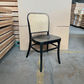 A black metal dining chair with a woven cane backrest, positioned in a warehouse setting with stacks of wooden boards and cardboard boxes in the background.