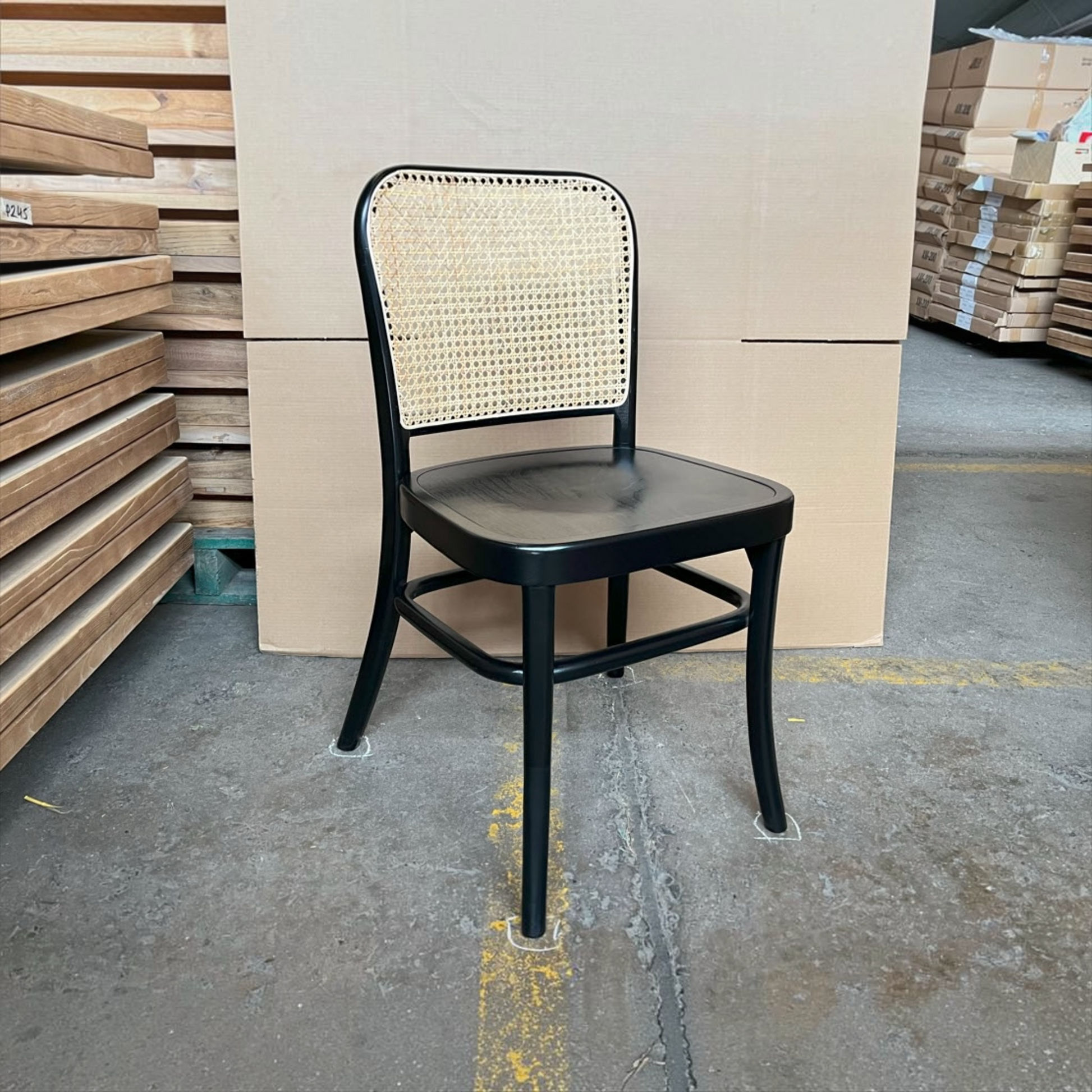 A black metal dining chair with a woven cane backrest, positioned in a warehouse setting with stacks of wooden boards and cardboard boxes in the background.