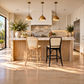 A modern kitchen island with a marble countertop, light wood cabinetry, three brass pendant lights, and two bar stools—a natural rattan cane chair and a black metal chair—with a marble seat. The room is brightly lit with warm sunlight and shadows on a light wood floor.