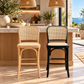 Two modern bar stools in a bright kitchen setting. The left stool has a light natural wood frame with a woven cane back and seat.