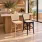 Two bar stools in a bright kitchen setting: one natural wood frame with woven cane back and seat, and one black metal frame with a woven cane back and seat. The scene includes a stone countertop, pendant light, plants, and large windows overlooking the sea.