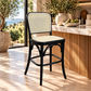 A black bentwood bar stool with a woven cane backrest and seat sits in a sunlit modern kitchen or bar area with marble countertops, potted plants, and a bowl of limes.