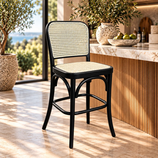 A black bentwood bar stool with a woven cane backrest and seat sits in a sunlit modern kitchen or bar area with marble countertops, potted plants, and a bowl of limes.