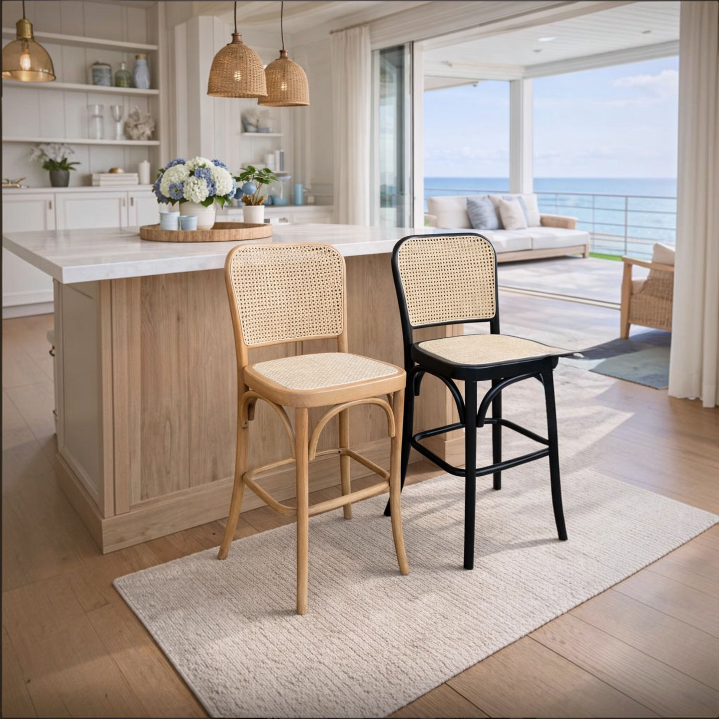 Two bar stools in a bright, modern kitchen with an ocean view. One stool has a light natural wood frame and woven cane seat, and the other has a black metal frame with a woven cane seat.