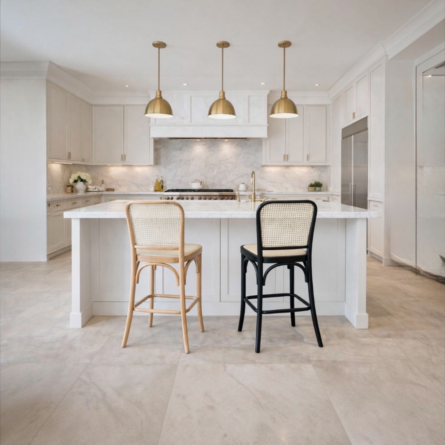 A modern kitchen island with a white marble-look countertop and white cabinetry, featuring two bar stools in front: one natural wood cane-back stool and one black cane-back stool. Three gold pendant lights hang above, and the room has light stone flooring and backsplash.