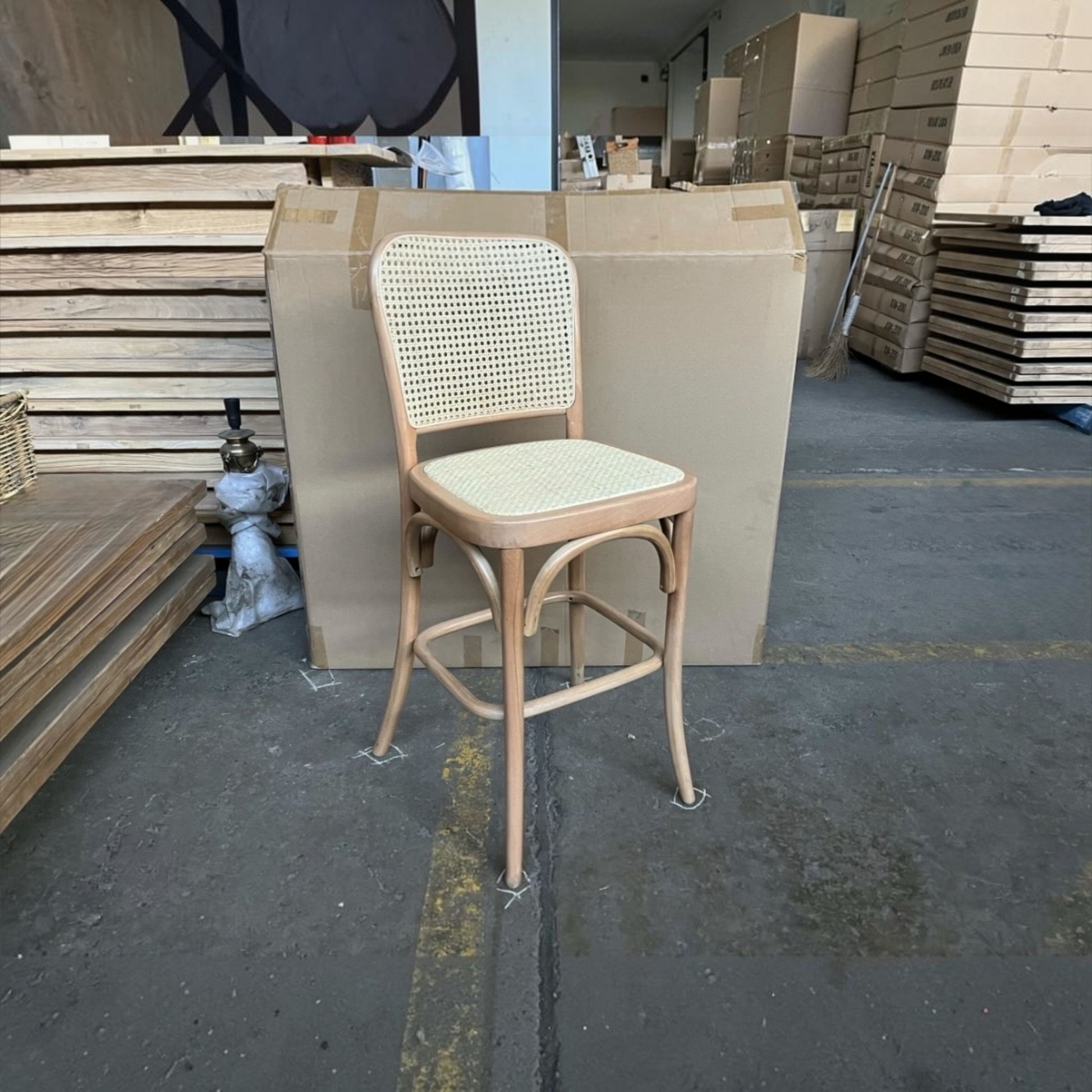 A wooden bar stool with a woven cane backrest and seat, positioned in a warehouse setting with large cardboard boxes in the background.