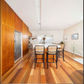 A modern kitchen and dining area with warm wood cabinetry, a white island, and four black bar stools with woven seats, set on a polished wood floor.