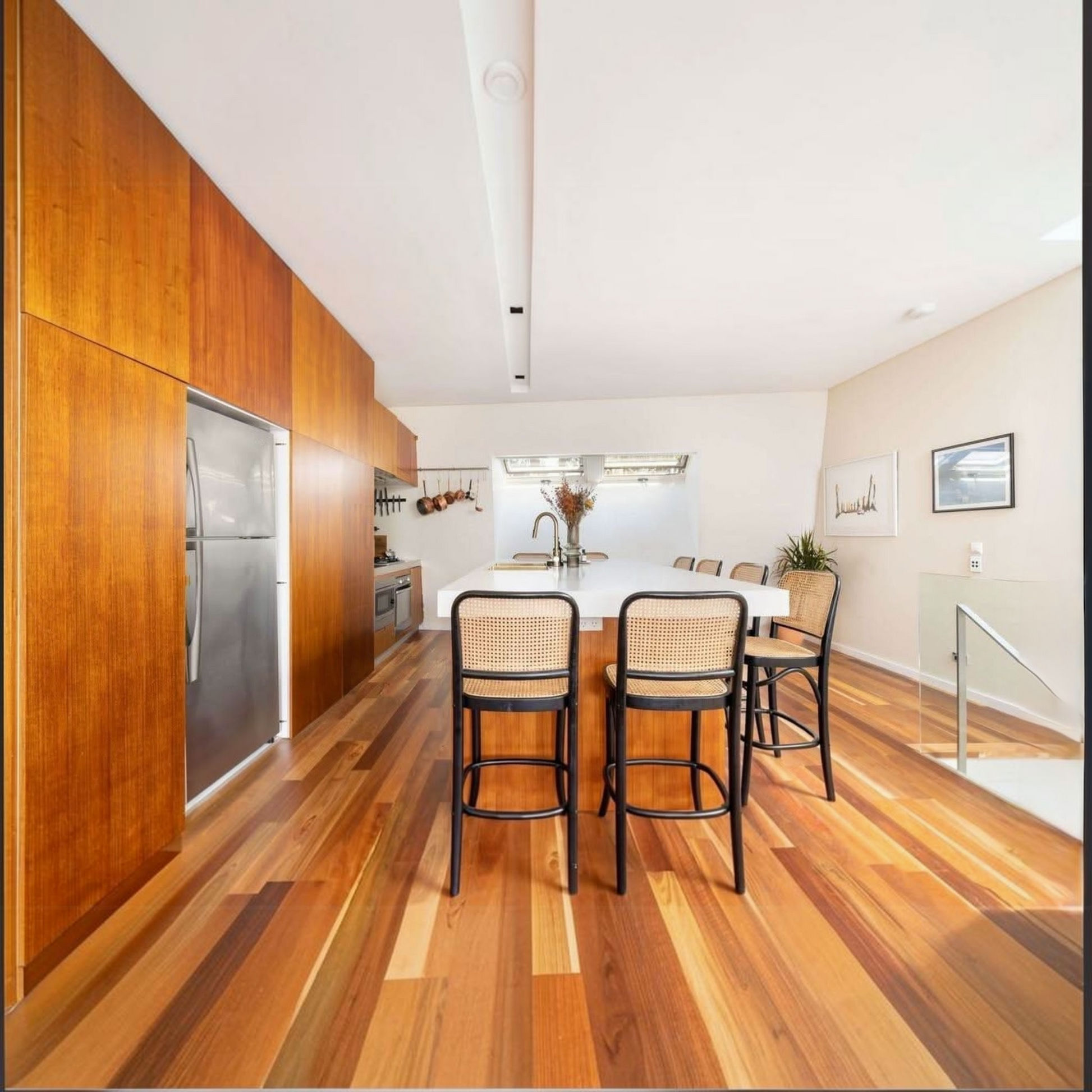 A modern kitchen and dining area with warm wood cabinetry, a white island, and four black bar stools with woven seats, set on a polished wood floor.